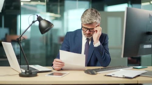 Serious mature businessman in a formal suit examines a document sitting at work in office Gray haire