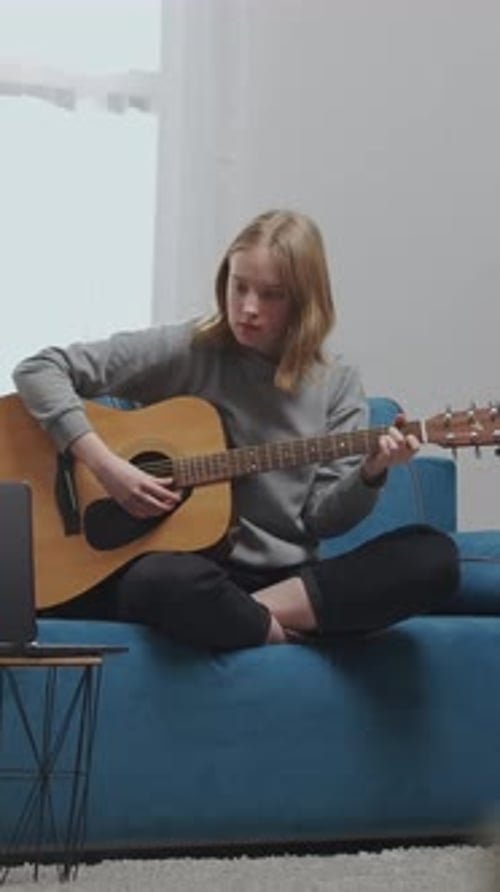 Teen Girl Practicing Acoustic Guitar on Blue Sofa at Home Focusing on Chords and Fingerpicking