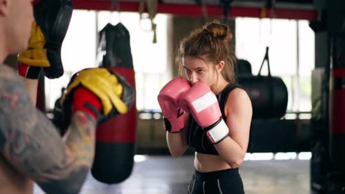 in a Boxing Fight Club a Young Girl in a Black Sweatshirt with Pink Gloves Practices with a Trainer