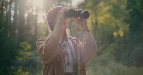Femme portant un chapeau en tricot regardant à travers des jumelles dans la forêt