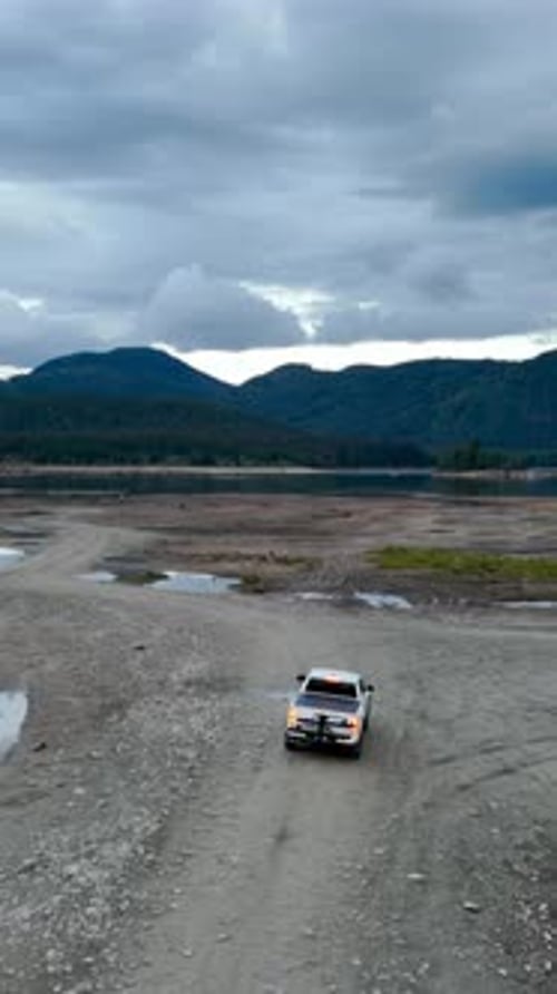 White Pickup Truck Driving on a Dirt Road Towards a Serene Lake and Majestic Mountains