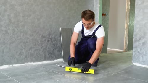Worker Sitting on Floor and Installing Carpenter Level on Freshly Laid Tiles in Apartment