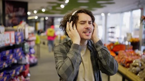Happy Brunette Guy with Curly Hair and Headset Listens to Cheerful Music While Shopping in a Grocery
