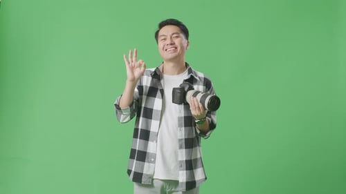 Asiatischer Fotograf schaut sich die Bilder in der Kamera an und zeigt eine Okay-Geste auf dem grünen Bildschirm