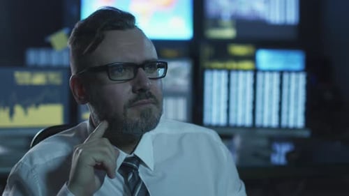 Manager is working on a computer in the evening in a dark office room filled with display screens.