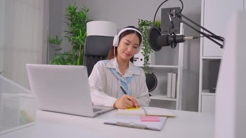 Asian podcaster in headphones using microphone broadcasting in studio.