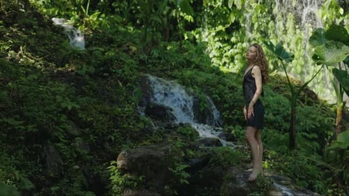 Charmante ruhige Dame steht in schwarzem Kleid vor dem Hintergrund eines kleinen Wasserfalls und genießt den