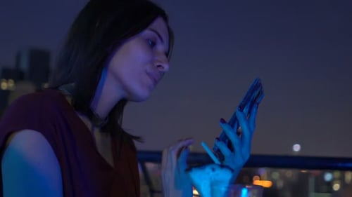 Young woman browsing internet on smartphone at rooftop bar terrace at night