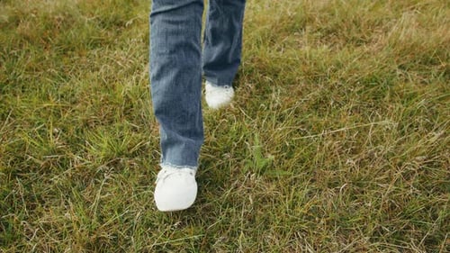 Young Woman Wearing Blue Jeans and White Sneakers Walking on Green Grass at Sunny Day Closeup Walk