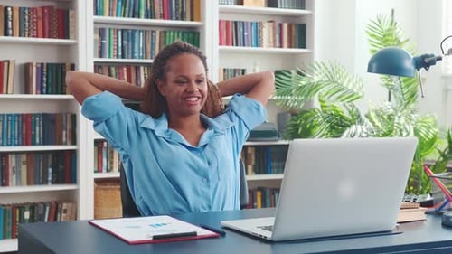 Young African American Woman Leans Back Sits at Table with Laptop in Office