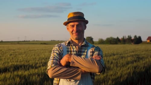 Portrait of smiling satisfied farmer whit hat standing in wheat field at sunset looking at camera.