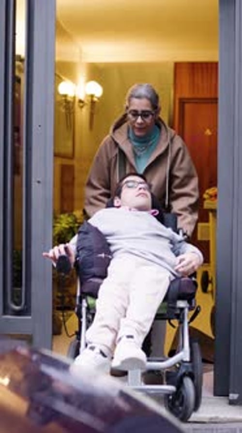 Mature Caregiver Pushing Young Man in Wheelchair Out of a Building