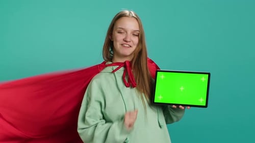 Joyeuse fille représentant un super-héros avec une cape rouge présentant une tablette à écran vert