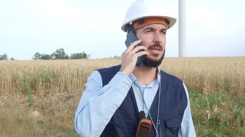 Male Engineer with a Safety Helmet Talking on the Phone in a Field of Windmills