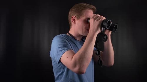 Man Looks Through Binoculars, Stands Against Black Background