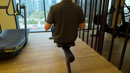 Man Exercising On A Gym Rowing While Looking Out A Window Urban Onto A City Street.