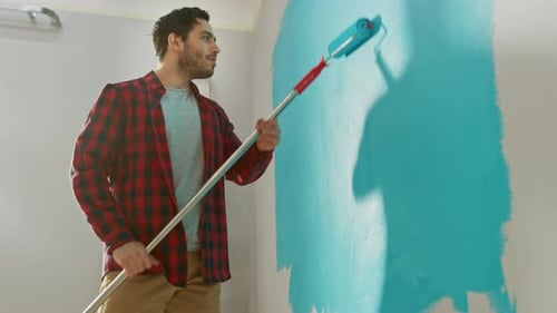 Mann in braunen Jeans und rotem kariertem Hemd malt eine Wand mit einer Rolle. Die Lackfarbe ist hellblau
