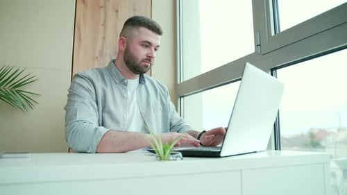 Concentrated young man freelancer working from the office sitting by the window, at the laptop,