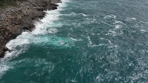 A Drone Flies Along a Rocky Ocean Coast Against Which a Strong Sea Wave of Bright Blue Color Breaks