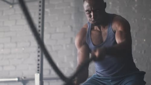 Using battle ropes, muscular man exercising intensely in gym, focusing on workout