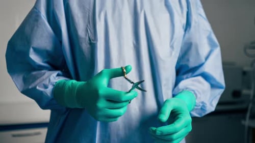 Closeup in an Operating Room Wearing a Blue Gown and Green Gloves Twirling Scissors in His Hand