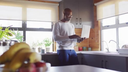 Happy senior african american man in log cabin, standing in kitchen and using laptop, slow motion