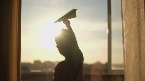 A Child Plays with a Paper Plane Near the Window of the House During Sunset