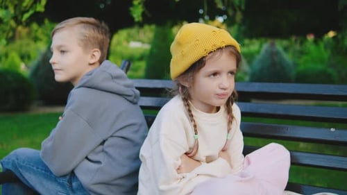 Zwei Kinder sitzen mit dem Rücken zueinander auf einer Parkbank, eines wirkt verärgert, während das andere