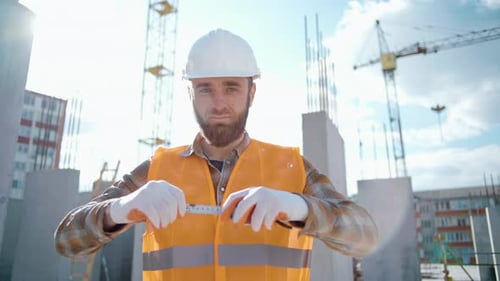 Young handsome builder in a hardhat holds a tape measure ruler in his hands and looking at camera