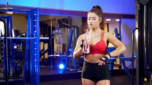 portrait of young attractive female bodybuilder drinking water from bottle in sports club indoors.