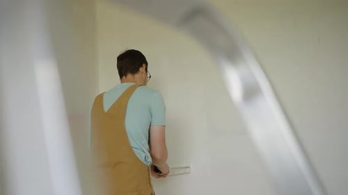 Professioneller Maler, der weiße Farbe von hinter der Leiter auf die Wand aufträgt und einen braunen Overall trägt und