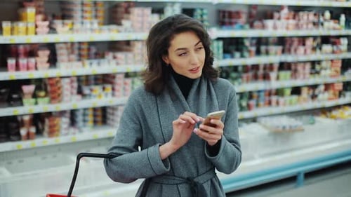 Young Woman Walk Checking to Do List on Phone in Supermarket Smiling Thinking Shopping Internet Buy