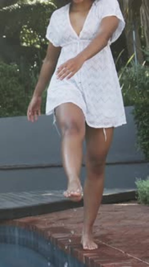 Vertical video: Reaching woman in white dress dipping toes on brick edge, walking poolside relaxing
