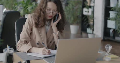 Asian Office Worker Speaking on Mobile Phone Writing Information in Notebook and Using Laptop