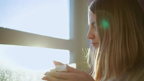 Side View of a Young Blonde Woman Sitting Near Window in Soft Sunlight