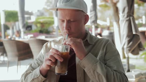 A Young Man is Sitting in a Cafe in the Fresh Air and Drinking Iced Coffee