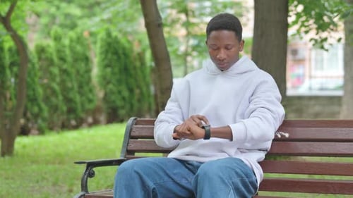 Young African Man Using Smart Watch on Park Bench