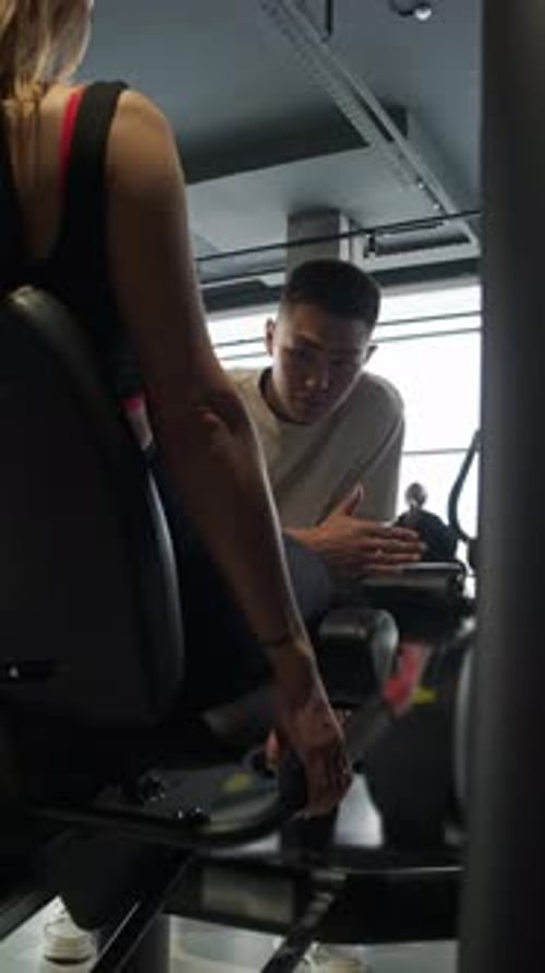 Young Woman Under Guidance of Personal Trainer Performs Exercises on Leg Simulator in Fitness Center