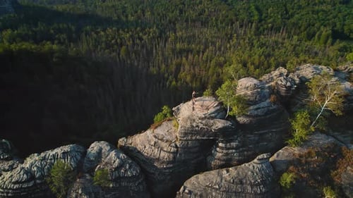 Hiker looks out over dense forests and distant farmlands, framed by rocky outcrops. A lone figure st