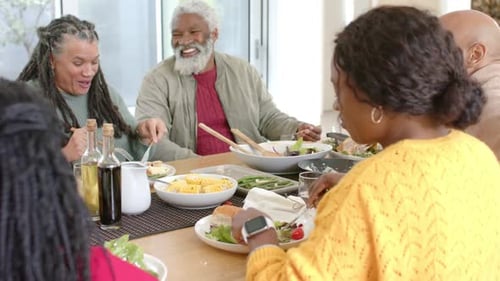 Rindo, uma família extensa diversificada está distribuindo saladas e acompanhamentos pela mesa de jantar em casa para jantar