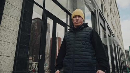 Person with a Focused Expression Wearing a Yellow Beanie and Black Puffer Vest Walking in Front of a