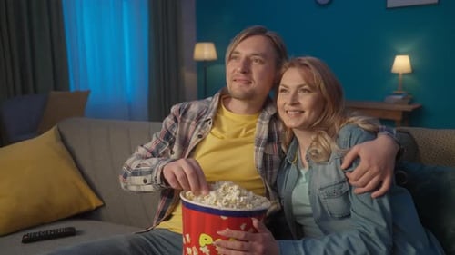 A Couple is Watching a Movie Eating Popcorn Man and Woman Hugging Relaxing on a Sofa in the Living
