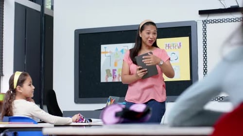 Teacher in classroom holding tablet, smiling at students during school lesson