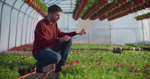 Man Inspecting Blossoms and Taking Notes in Greenhouse