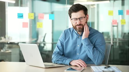 Upset businessman suffering from toothache working on laptop sitting at workplace in business office