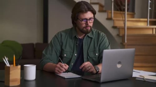 Man Writing in Notebook with Laptop at Home