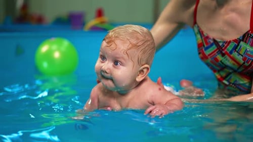 Adorable blond baby boy held by his mom in the water of swimming pool.