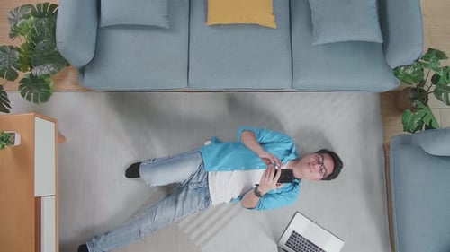 Draufsicht Zoom-Out-Aufnahme eines asiatischen Mannes, der auf dem Boden liegt und das Smartphone zu Hause benutzt