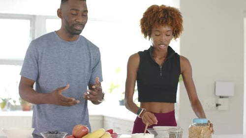 Diverse couple in kitchen, gray tee man gesturing, woman in black crop topping bowl with seeds