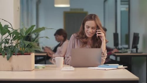 Smiling business woman working on laptop and chatting mobile phone in modern office